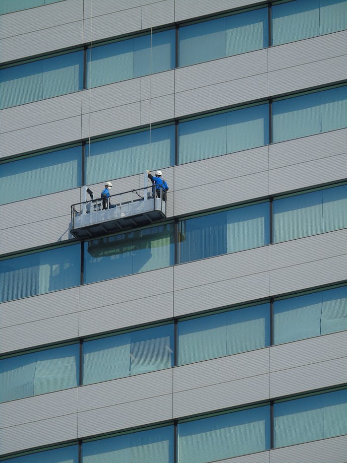 Two workers clean windows on a modern glass building facade using a suspended platform.